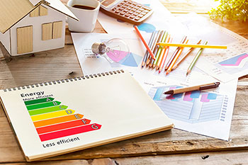 Diagramma classi energetiche su un blocco a spirale appoggiato su un tavolo con matite colorate, altri fogli con diagrammi vari, una calcolatrice e un modellino in legno di una casa.