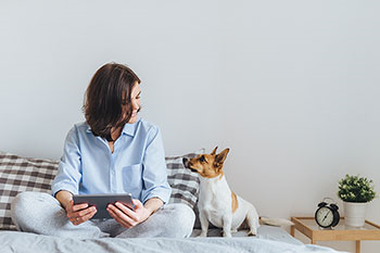 Giovane donna seduta sul letto con un tablet in mano guarda il suo cane che la guarda