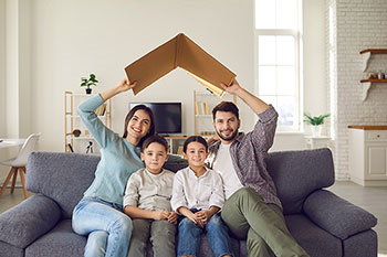 Famiglia seduta sul divano, i genitori tengono sopra le teste un pezzo di cartone a simulare il tetto di una casa