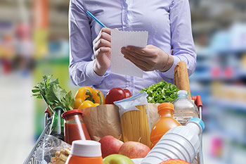 Donna con foglio e penna in mano scrive davanti a un carrello della spesa pieno di prodotti; sullo sfondo, sfocato, l'interno di un supermercato.