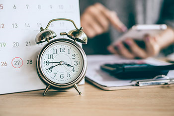 Calendario da tavolo con data di scadenza e sveglia, con sullo sfondo mani di donna calcolano le spese mensili con una calcolatrice.